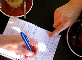 Aerial view of a cafe table, showing two empty coffee cups, a notebook page of notes, with one person pointing to it and the other making notations.