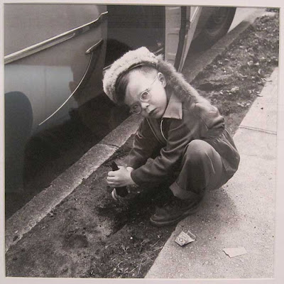 Young boy in coonskin cap, squatting beside a car, digging in the dirt, looking up at the photographer almost angrily