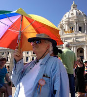 Silver-haired man with rainbow umbrella standing before the Minnesota State Capitols dome and gold horses