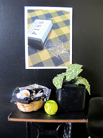 Detail of a modern dolls' house miniature scene of a vintage sewing table and Eames chair in front of a black wall displaying a poster with vintage pins on a checked fabric.