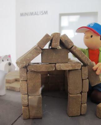 Miniature clay figure, building a dog house out of bricks in a gallery, with a dog looking on.
