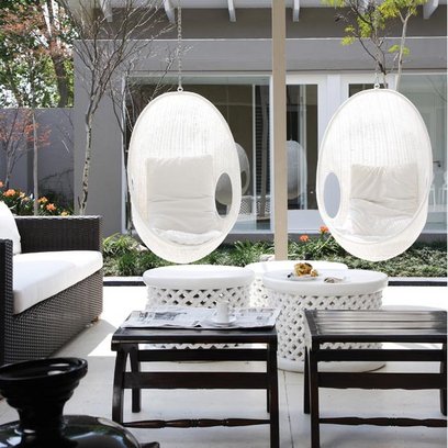 White hanging chairs in a family room with glass walls and dark wicker sofa with white cushions, two dark wood side tables and round white footrests with woven detailing on the side
