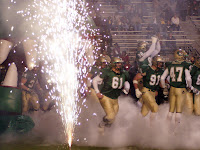 football players in green and yellow uniforms take the field amongst fireworks and smoke.