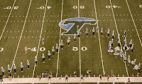 Marching band takes a football field and forms a wave formation at the fifty yard line around a T with a wave drawn over it at midfield.