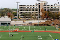 Aerial view of a college football practice facility with a track around the field and construction of tall building in the background.