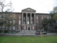 Three story wide rectangular building with four distinct columns. Stairs leading up to the majestic dated brick structure merge from green lawn in front of the building.