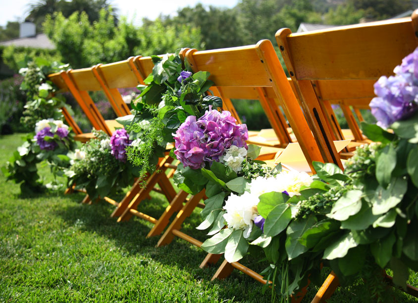 A garland with purple hydrangea is draped on the backs of the chairs at a 