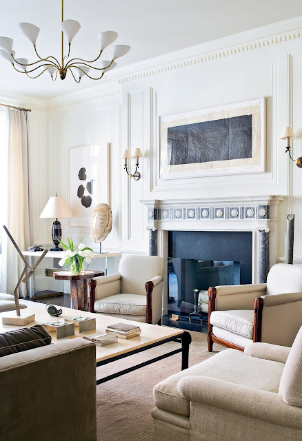 Living room by Peter Pennoyer with decorative moulding, a gold and white chandelier, fireplace with blue and white marble detailing, white armchairs, a taupe sofa, cream are rug, a large coffee table and wood side table