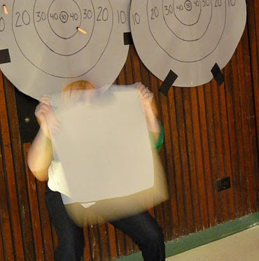 Image of a jokey/nervous helper hiding behind a vecro vest from a Nerf Foam Bullet barrage, triggered by people using accessibility switches to fire at her.