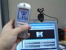 this is a pic of a laptop and a webcam, there is also a white hand in the pic holding up a cell phone