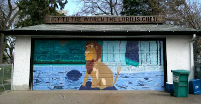 Surburban garage door painted with a mural of an adult Simba with Nala. On the roof of the garage is a wooden sign that says Joy to the World the Lord Is Come