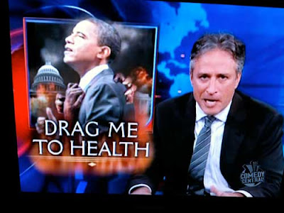 John Stewart, with graphic showing Obama, type labeling it Drag Me to Health