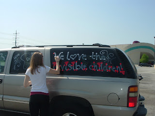 Young lady decorating side of a beige SUV with words ''We love the Invisible Children''