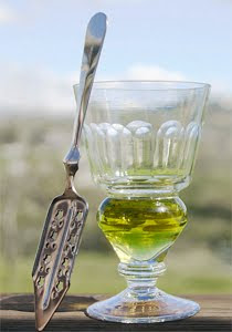 A reservoir glass filled with a naturally colored verte absinthe next to an absinthe spoon.