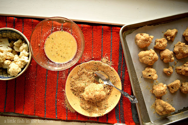 A small assembly line for making breaded cauliflower. A bowl with fresh cut cauliflower florets is first in line, next to a bowl of egg wash, and finally another small shallow bowl, half full with bread crumbs. A single cauliflower floret is in the center of the shallow bowl and is covered in bread crumbs. A metal tablespoon, used to coat the veggies, rests in the shallow plate too. At the end of the line, there is a metal sheet pan, covered with parchment paper and half full with breaded cauliflower to be baked. 