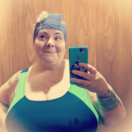 image of me in a blue and green swimsuit and a retro purple swimcap with flowers, standing in a mirror grinning with my eyes looking to one side