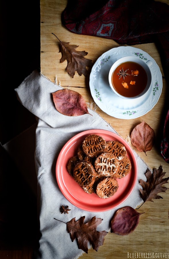 Vista cenital, seevilleta vintaje burdeos, taza porcelana antigua con té, Galletas caseras sobre plato cerámica rojo, fondo madera oscuro, manzanas rojas, hojas secas otoño, paño de lino beige