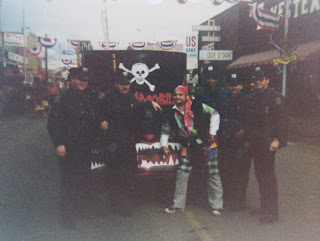 A color photograph of John Belushi and five men in police uniforms posing in front of a vehicle in the middle of a highly decorated street.