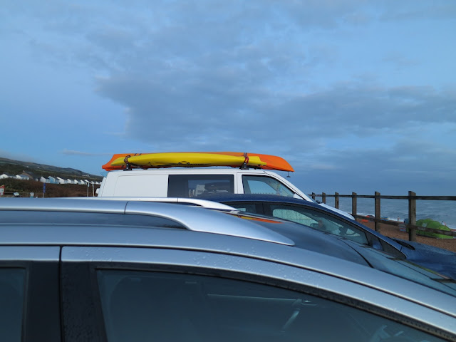 Orange and yellow canoe on the roof of a white van / car facing railings and sea