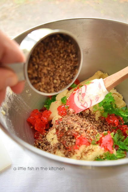 Basic sugar cookie dough, chopped candied cherries, finely chopped pecans are layered in side a metal mixing bowl. They are ready to be combined using a stand mixer.