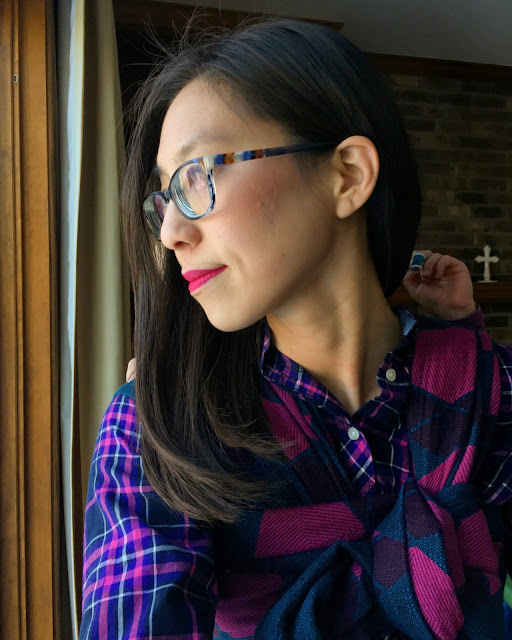 Image of a tan skin bespectacled Asian woman with dark brown hair looking out a window while wearing a child on her back in a pink and blue large scale argyle patterned woven wrap carrier over a navy, purple, and pink plaid top.