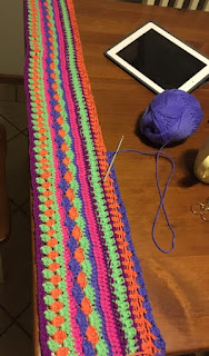 The edge of Sarah's blanket lined up across the edge of the table.  Also on the table is an iPad and a skein of purple-blue yarn which is the current colour being worked.