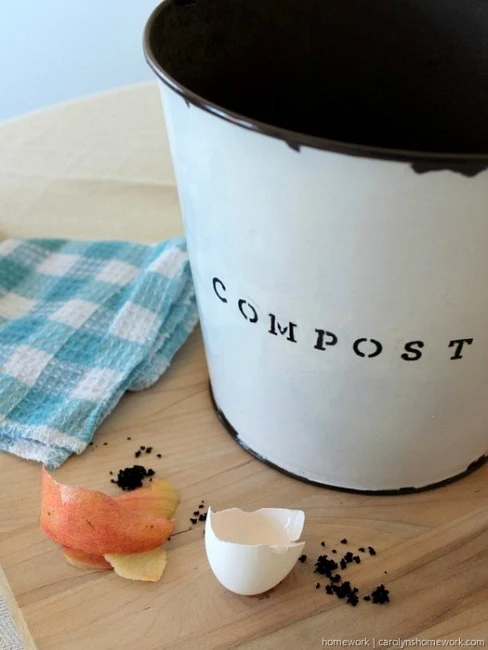 White painted galvanized bucket with the stenciled word compost on it, next to egg shells and peels and a blue checkered dish towel.