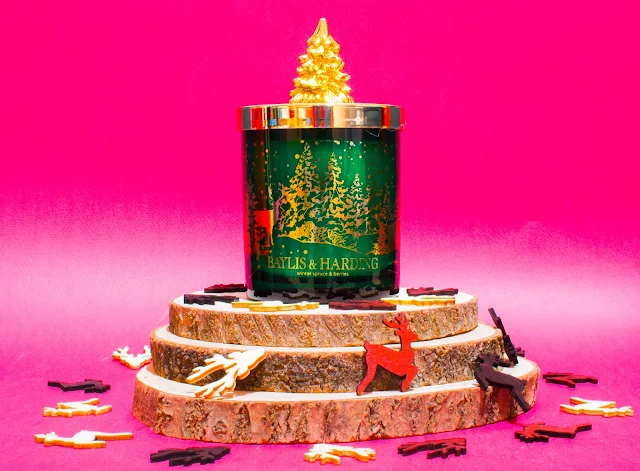 A candle in a green glass sjar with gold trees on and gold lid with a tree on and sitting on cut wood rounds with christmassy wooden confetti