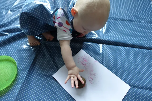 A baby making stamps with cut beetroot on a white piece of card on a wipe clean messy mat in blue with small white stars. Taken further away than the previous shot so you can see more of the baby.