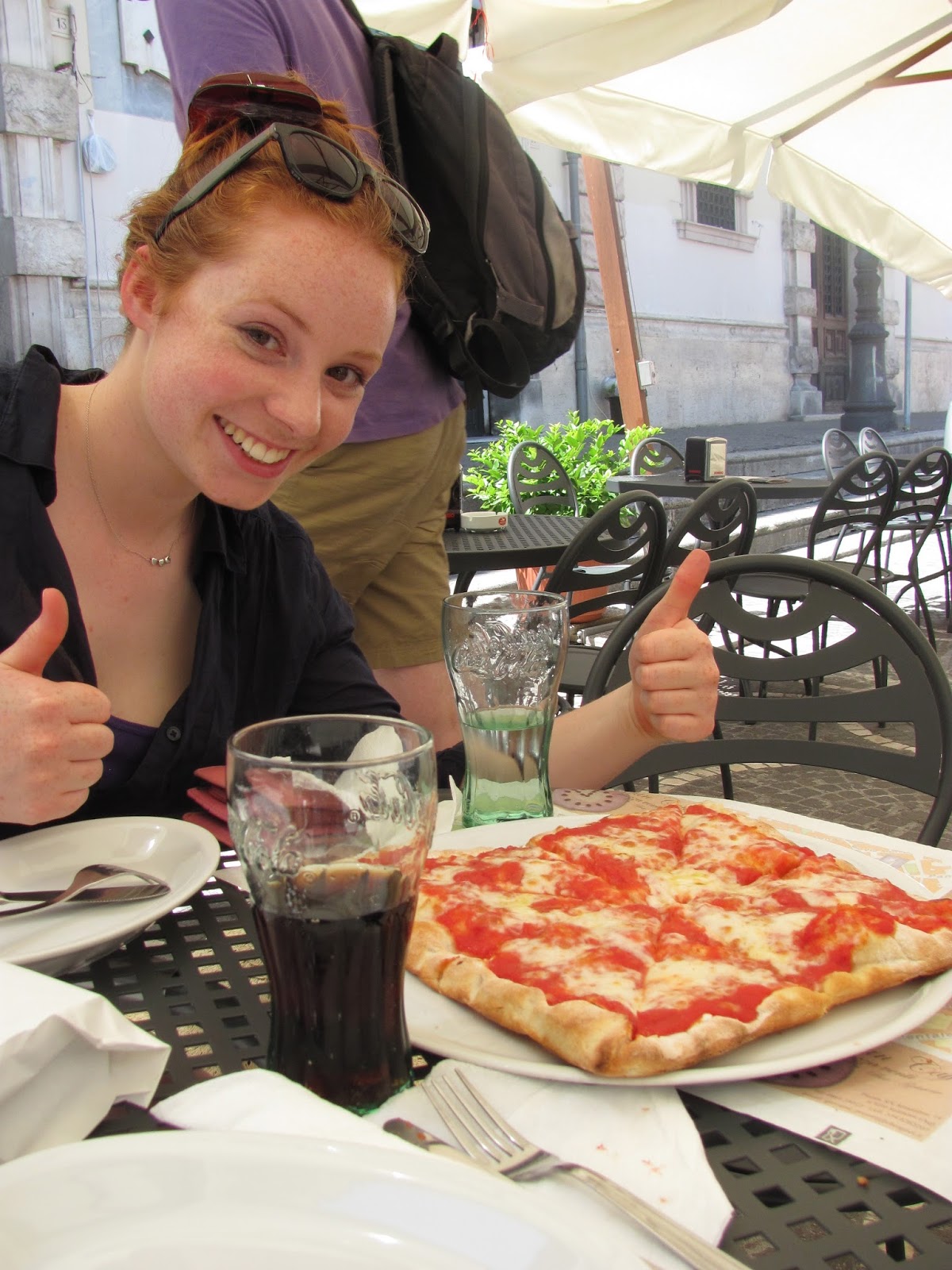 A young woman gives 2 thumbs up to a square pizza pie.