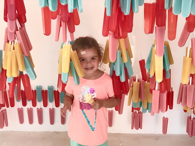 A toddler girl poses holding an ice cream cone in an art installation that has ice cream bars hanging from the ceiling