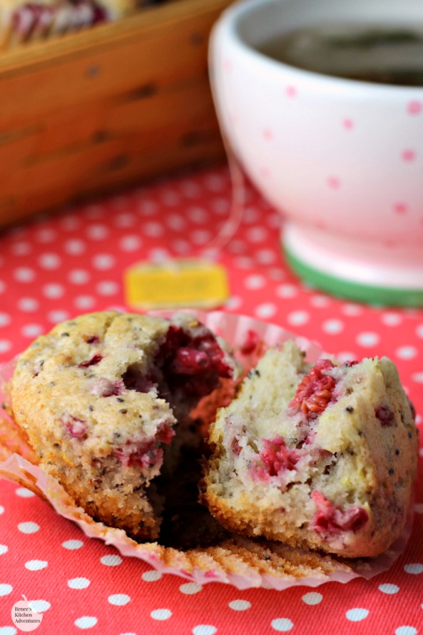 A Lemon Raspberry Chia Seed Muffin by Renee's Kitchen Adventures on a plate split in half to see what the inside looks like. A pot of tea and a basket of muffins in the background. 