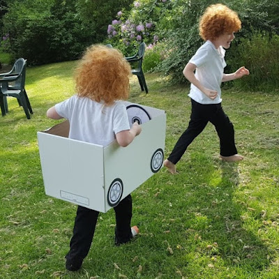 Two boys, one inside a cardboard car, chasing in a garden