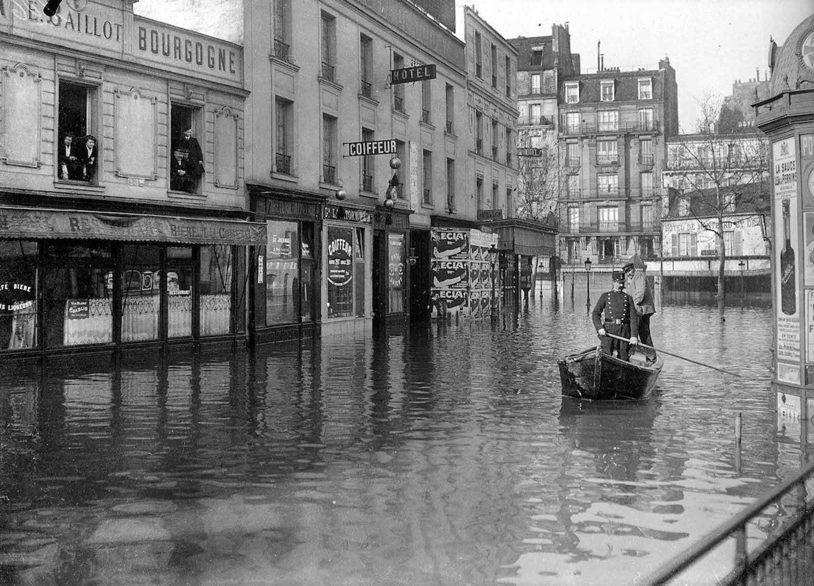 A uniformed man punts a boat as people at first floor windows look on.