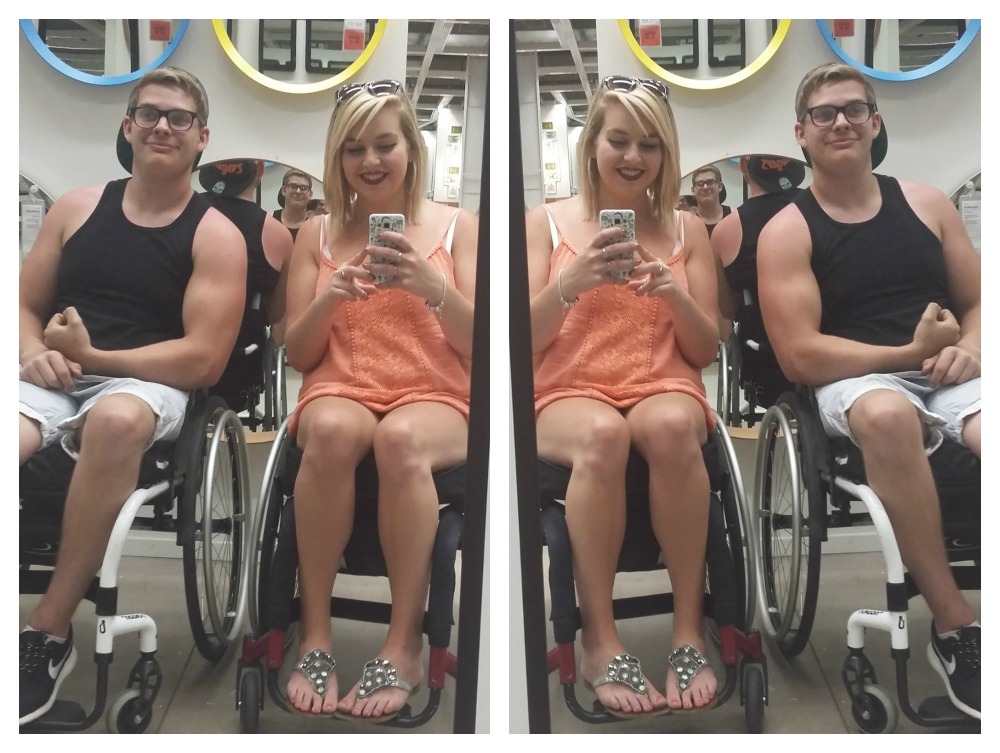 Image shows my brother on the left and me on the right. We are sat in wheelchairs and posing in a mirror in Ikea. Ollie is wearing a black vest top and shorts. I am wearing an orange vest top and shorts. There are more mirrors in the background