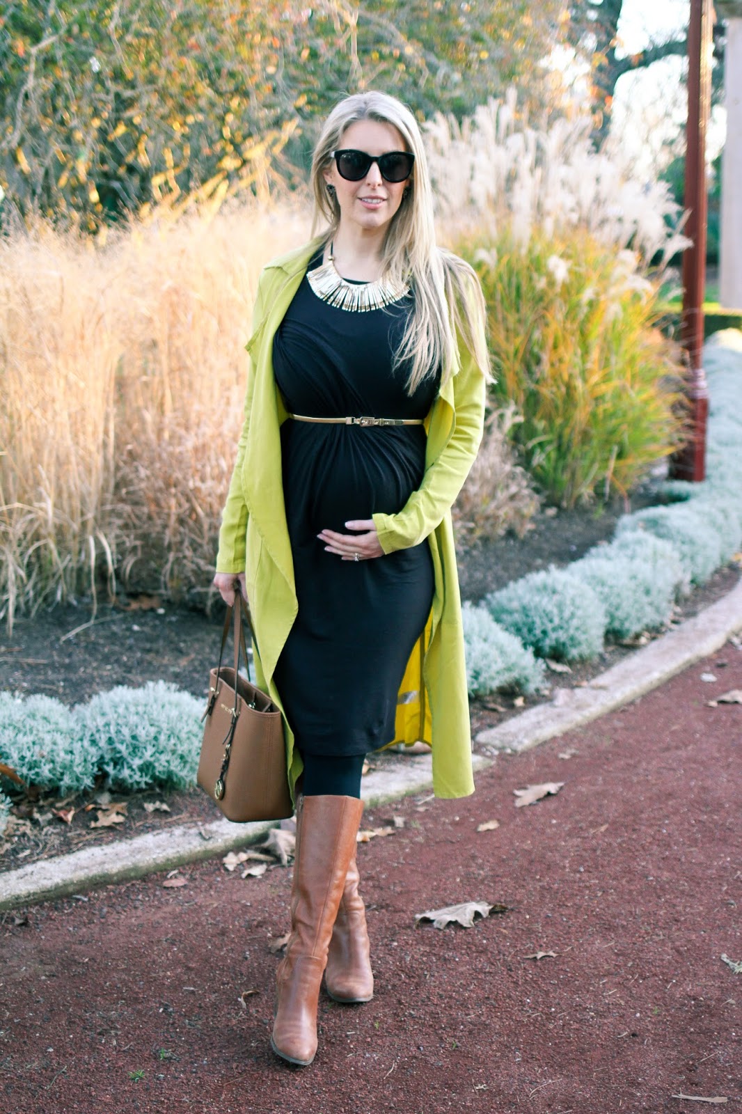 The Goldfields Girl styling maternity fashion. She wears black maternity dress, green jacket, statement gold necklace, brown boots and Michael Kors bag.