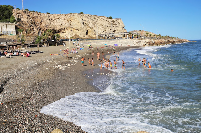 Pequeña playa de piedras y arena gruesa con gente en la orilla y el agua del mar con olas.