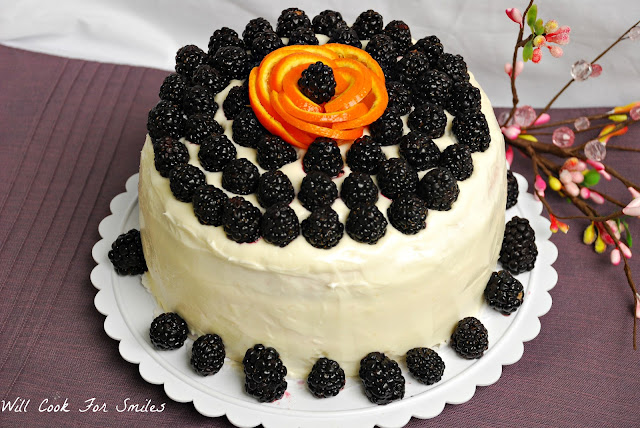 whole cake on a cake platter with white frosting and blackberries all around the bottom and blackberries all over the top with a orange peel made to look like a flower with a blackberry in the middle