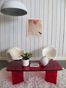 Modern dolls' house miniature scene with white paneled walls, concrete floor with a white flokati rug on top. Two white Eames chairs sit behind a red perspex coffee table and a large white light hangs overhead.