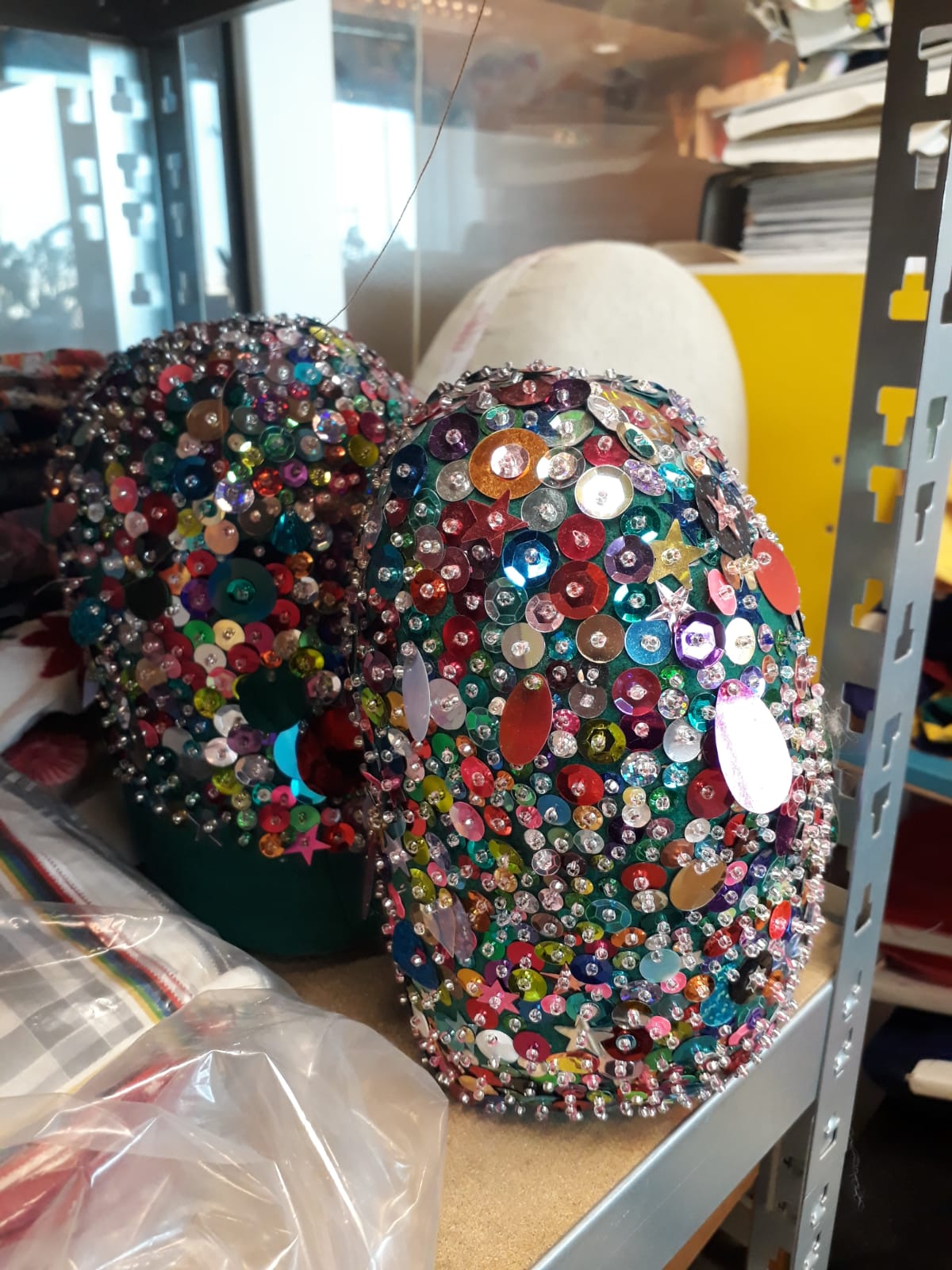 This picture shows a close-up of a shelf in Bas Kosters' studio. On it stands two small head-shaped pieces decorated with different sized and shaped sequins.
