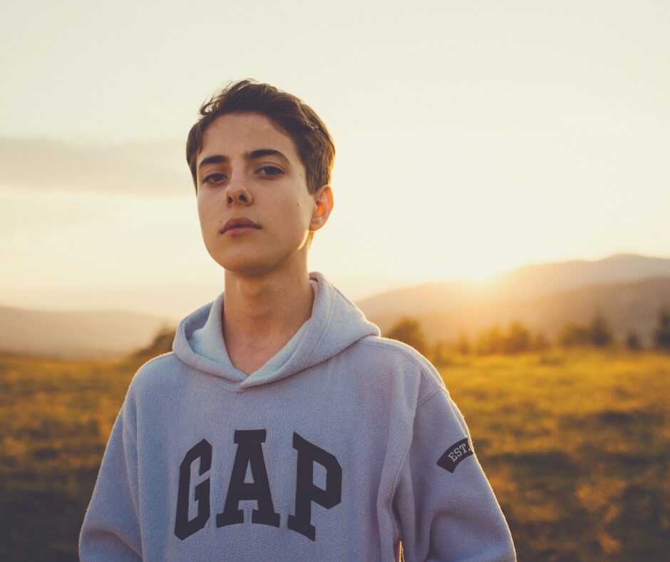 A moody teenage boy looks into the camera, no smile on his face. He is wearing a grey GAP hoodie, his eyes and hair are brown. The sun rises to the right of him in the background above  grassy mountains.