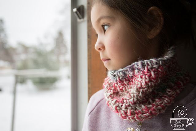 Image of a child looking out a door at the winter wonderland outside. Child has on a multi-colored cowl over a lavender sweatshirt.