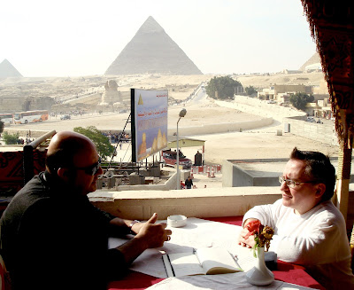 Simon sits opposite an Egyptian man in a coffee shop with the pyramids in the background.