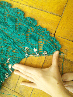 A shawl with a knitted-on leaf lace edging in progress. The edgiing is laying flat, and has been shaped by short rows to be at an angle to the body of the shawl. Numerous stitch-marker charms are clipped into the work. 