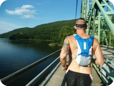 View of runner from behind wearing the CamelBak Marathoner Vest running across a bridge, with a mountain and river in the background