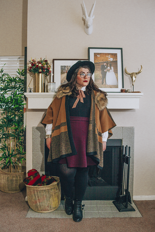 An outfit of a black wide brim fedora, brown faux fur collar poncho, peter pan collar blouse, black sleeveless dress, purple chevron print skater skirt, black tights and black heeled buckle boots.
