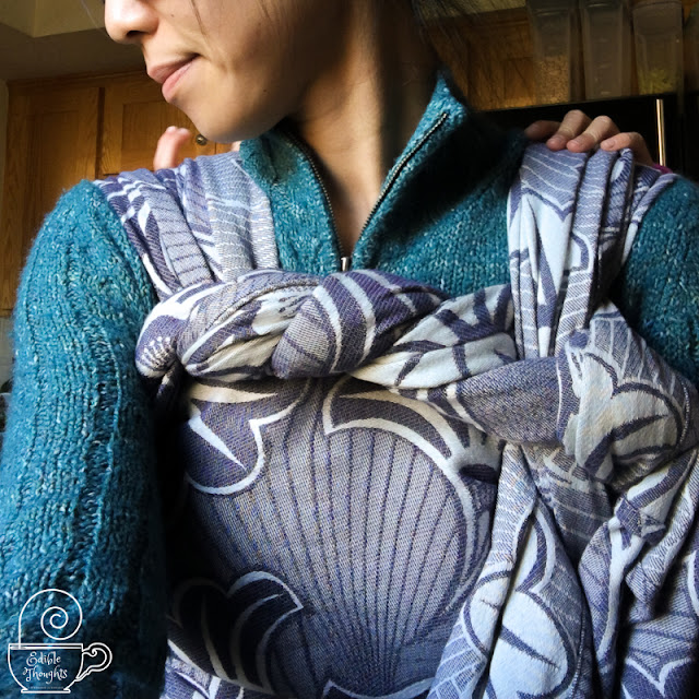 [Image of the upper torso from nose down of a tan skin Asian woman wearing a child on her back in a grayish-purple geofloral a woven wrap over a teal zip-up sweater. Child’s hand rests on woman’s left shoulder.]