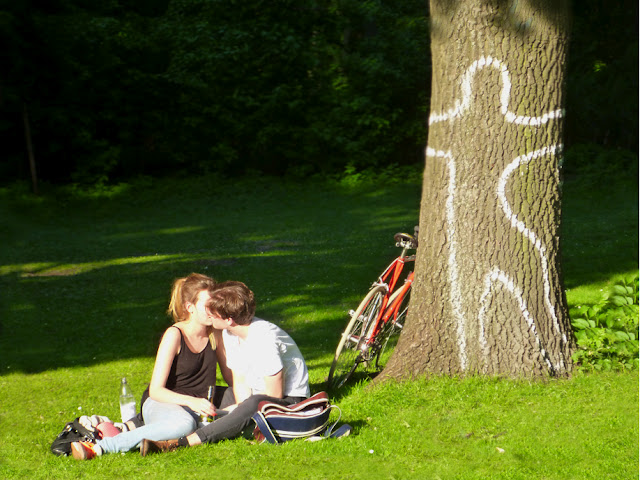 Ein Liebespärchen sitzt auf einer Wiese, hinter ihnen ein Baumstamm, auf den mit weißer Farbe der Umriss eines Menschen gemalt wurde. Ein Liebespärchen sitzt auf einer Wiese, hinter ihnen ein Baumstamm, auf den mit weißer Farbe der Umriss eines Menschen gemalt wurde.