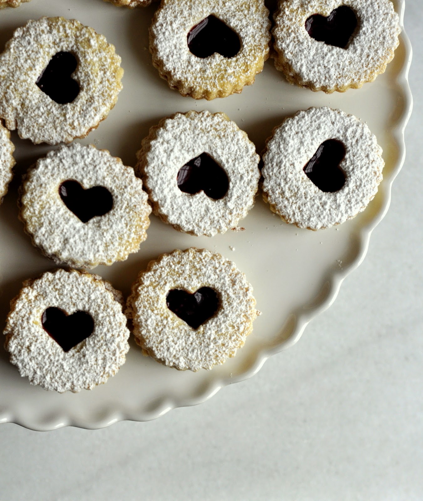 Raspberry Linzer Cookies with Heart Cutout Dusted with Powdered Sugar on White Cake Stand with Scalloped Edge | Taste As You Go