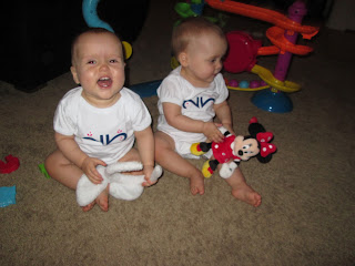 twin girls sitting on floor, one holding bunny ears one holding Minnie Mouse toy, both wearing white my twins are cuter onesies